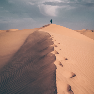 Desert trek individual An individual far in the distance walking across the top of a sand dune with foot prints behind them. Clouds gather in kt.