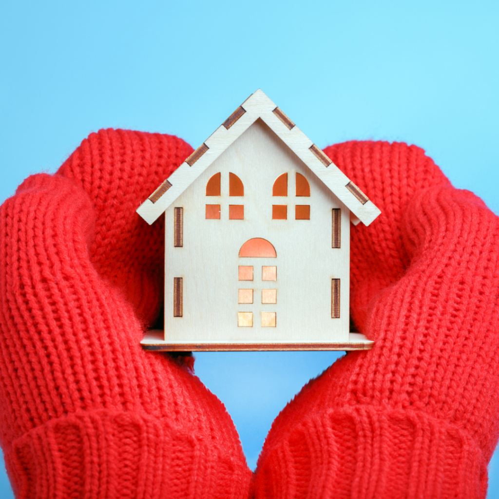 Two gloved hands holding a wooden model of a house what is glowing with warmth inside.