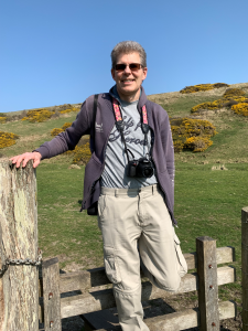 Martin Rickerby smiling and wearing a camera round his neck, standing infront of a stile in a fence, with a green hill and blue skies in the background.