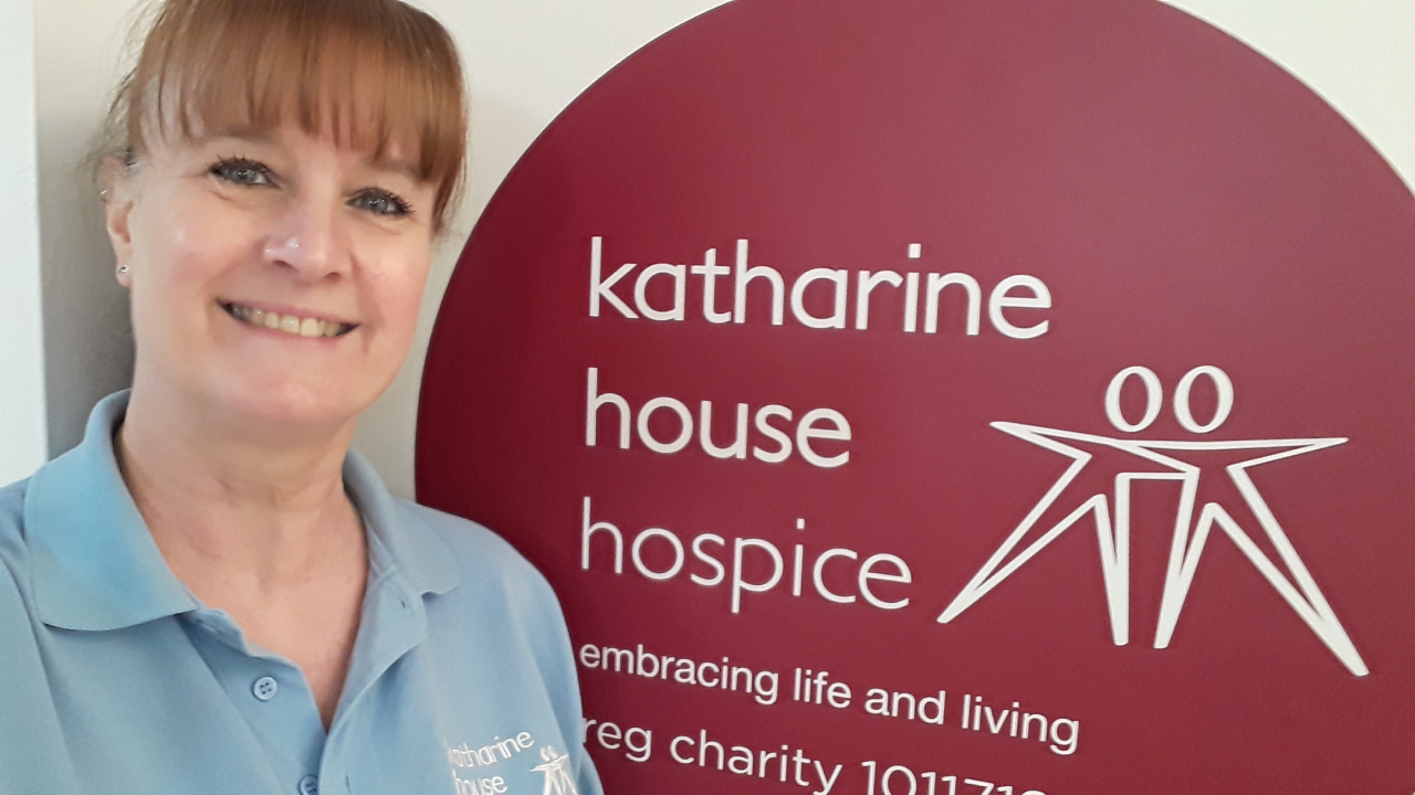 Sue Humpage, Health Care Assistant, standing next to the Katharine House Hospice logo.