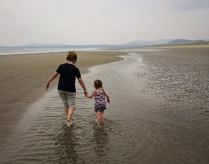 Woman and granddaughter on the beach walking away from the camera into the distance. They are holding hands and walking in a channel of shallow water on the sand.
