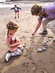 In the foreground, a young girl sitting on the beach making a heart symbol in the sand with stones. To the right, her grandmother helps her. In the background another young girl is playing.