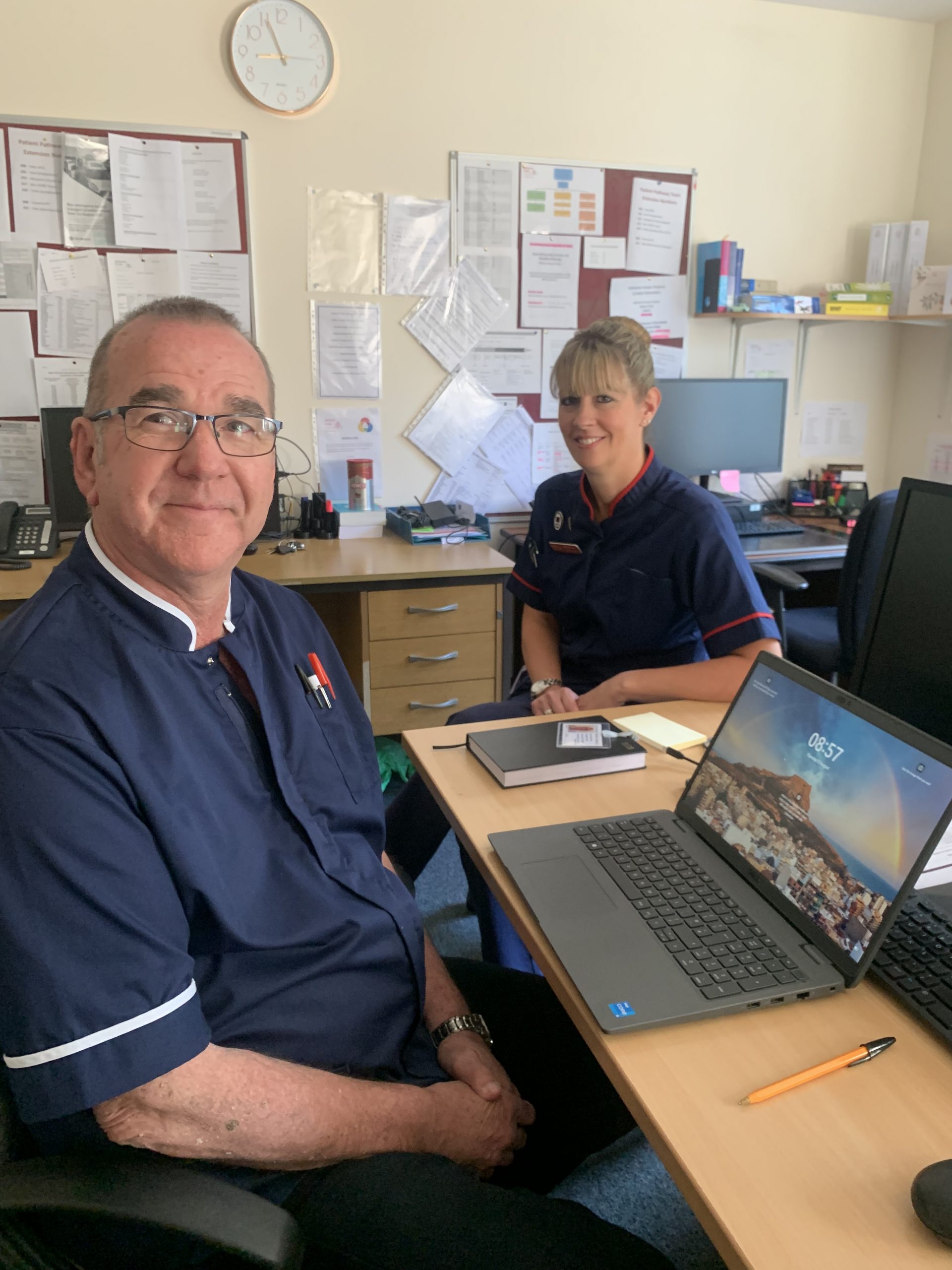 Palliative Care Nurses, Neil and Rachel, smile together at their desks.