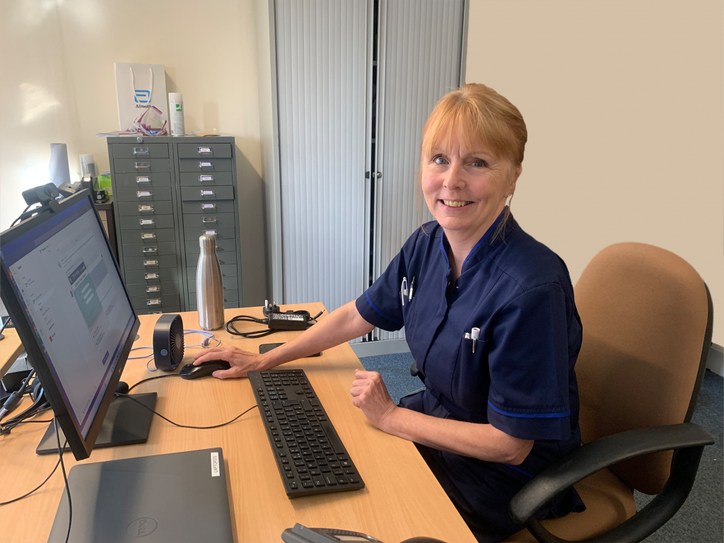 202308_Care_PCNS_Pat McGovern Palliative Nurse Pat sat at her desk and smiling to camera.