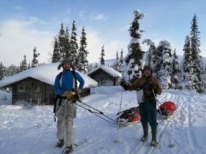 Two mean wearing cold weather gear and skis. The scene is one of a cold wilderness, featuring two wooden shelters.