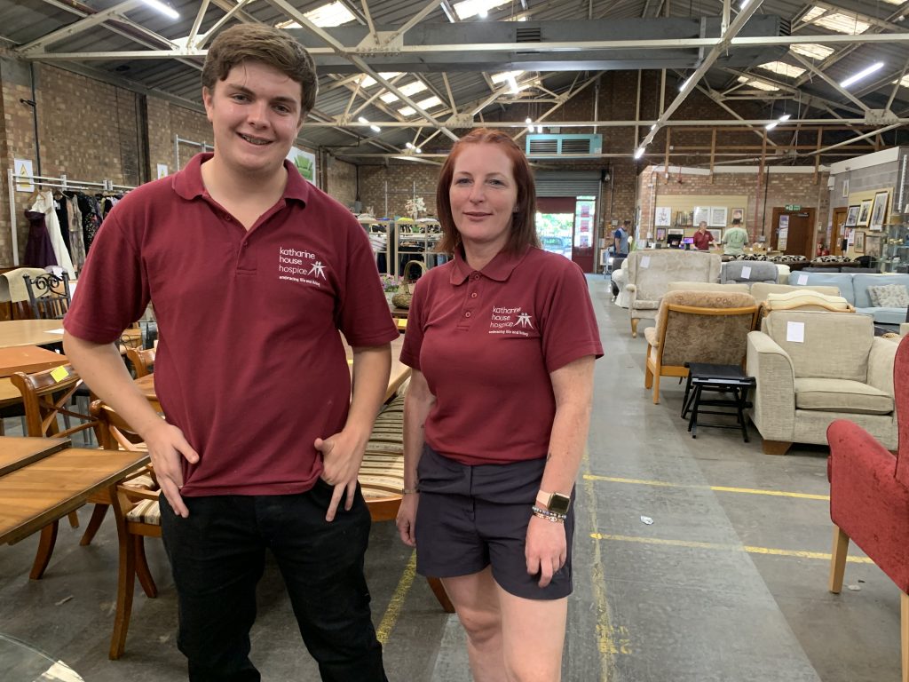 Two staff members stand in a warehouse charity shop surrounded by furniture