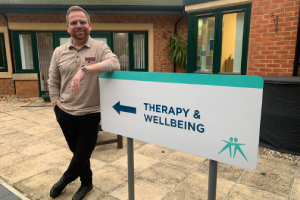 Javier, the family support team leader, stands next to our Therapy and Wellbeing sign, smiling to the camera.