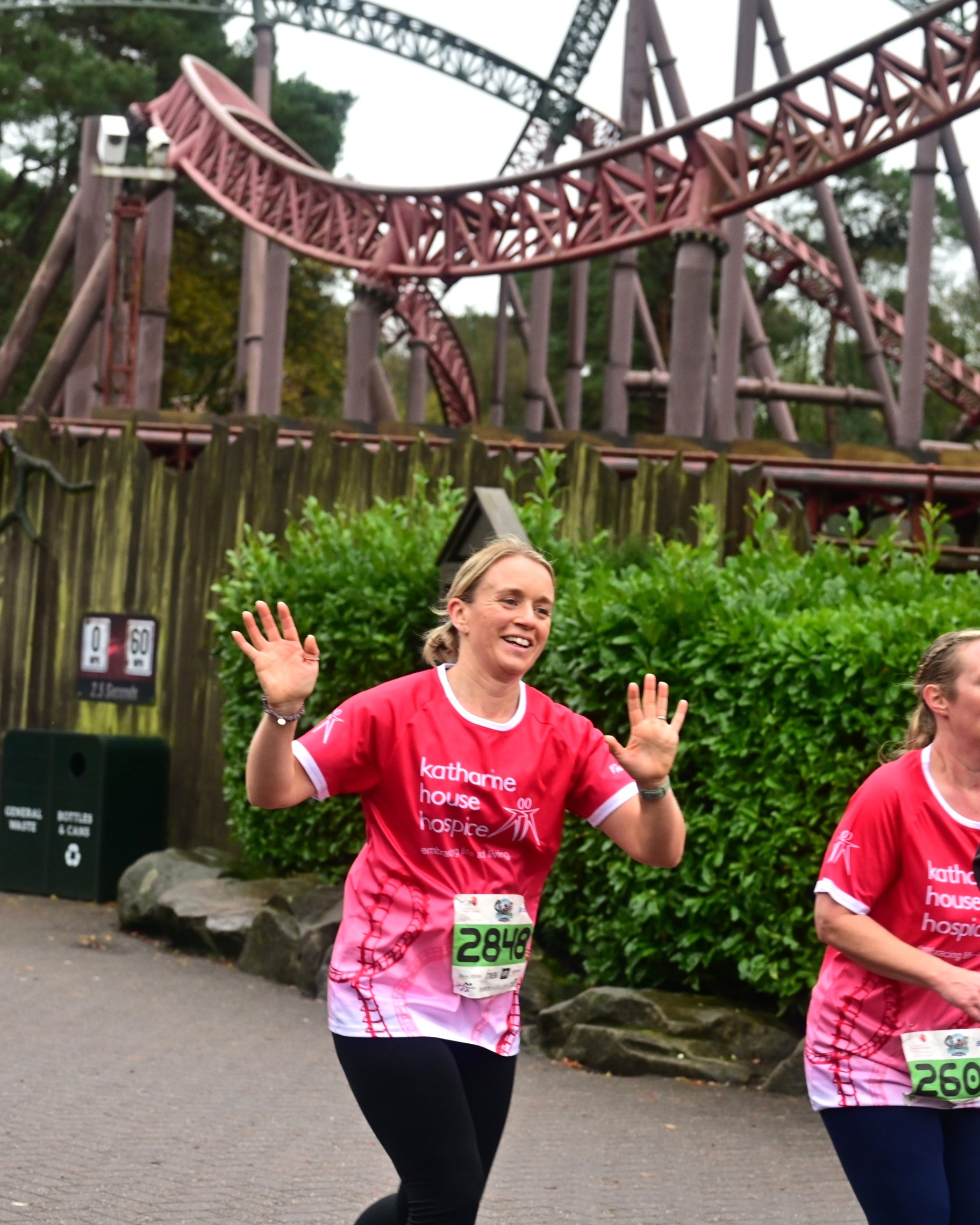 A woman in a charity run t-shirt smiles and waves while running past a roller coaster.
