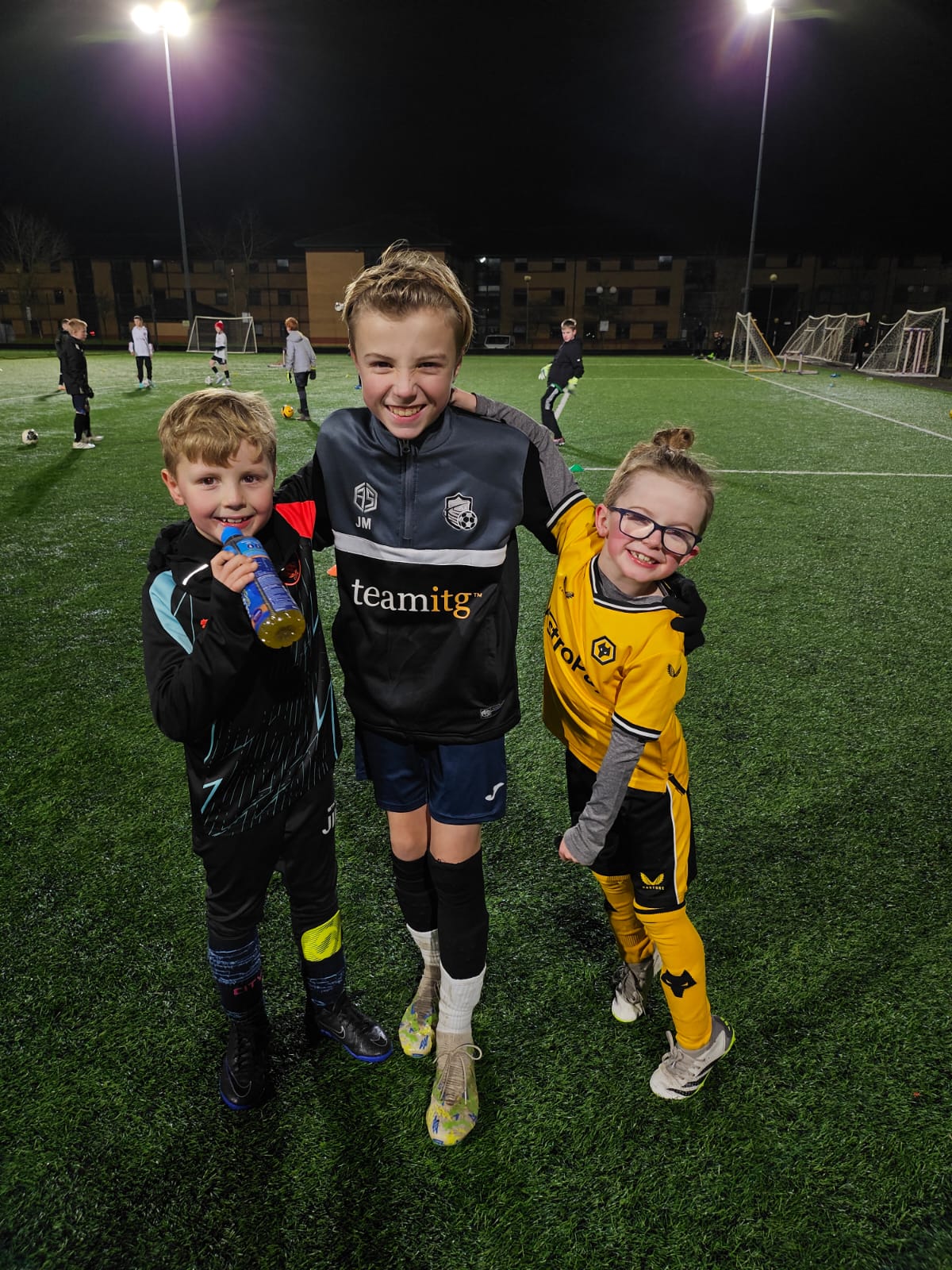 Three young boys stand smiling together on a football pitch