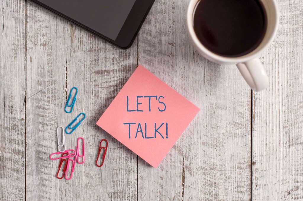 Let's talk Picture showing a coffee cup with black coffee and a pink note that says LET'S TALK on a wooden table top