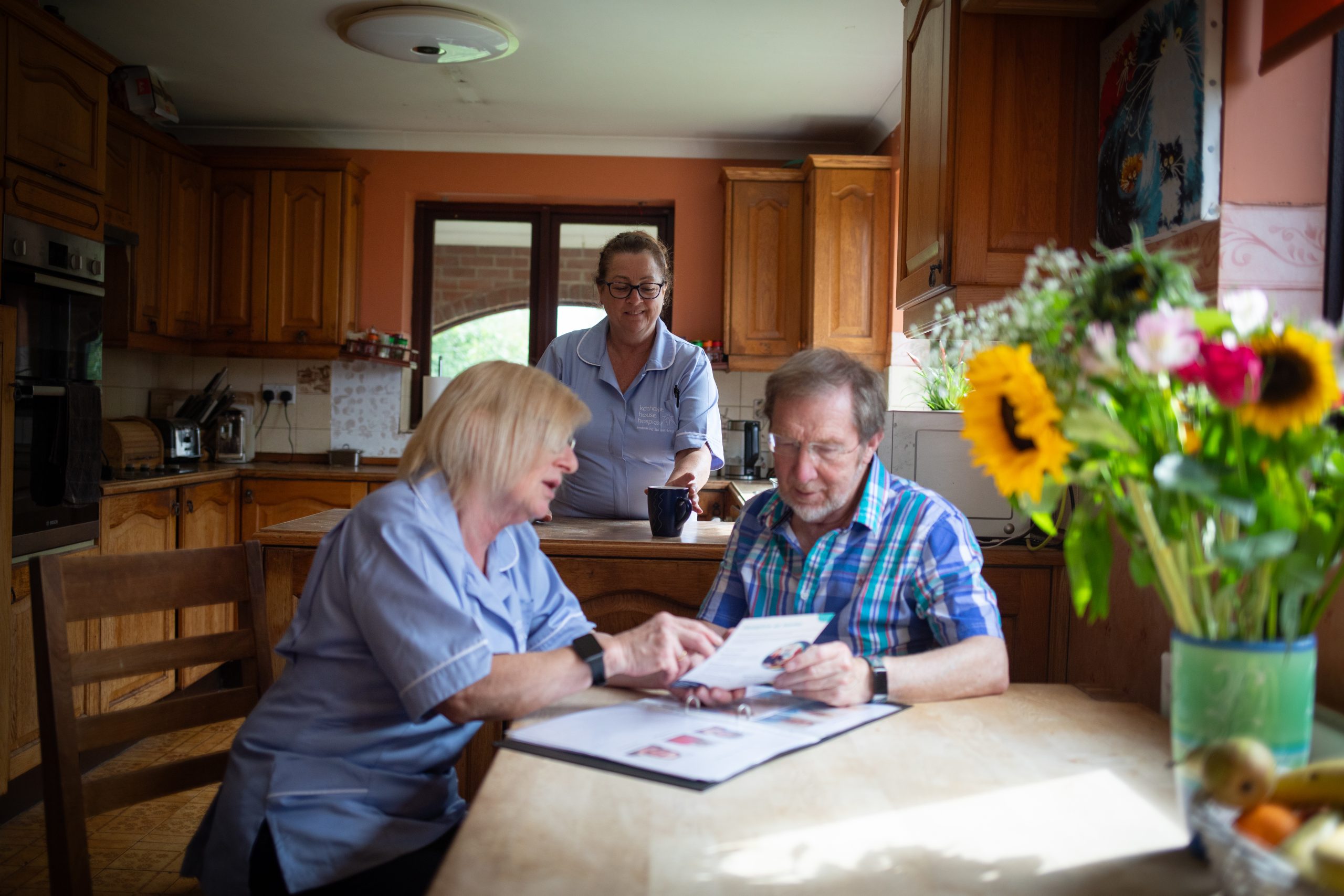 Hospice care at home (1) A nurse speaking to a patient in his home, while another nurse brings him a hot drink.
