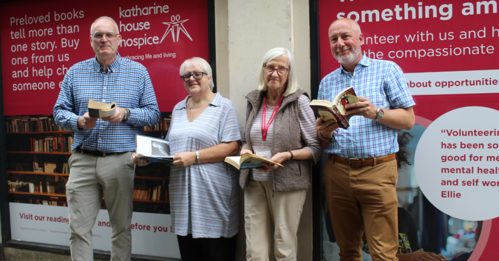 Four people standing outside a shop front all holding open books. The shop windows are filled with graphics promoting Katharine House Hospice retail.
