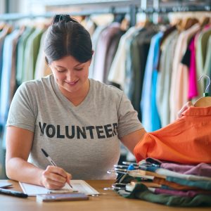 A young lady in a grey t-shirt that says 'volunteer', holding a pen and making notes on a counter with a pile of clothes on handers