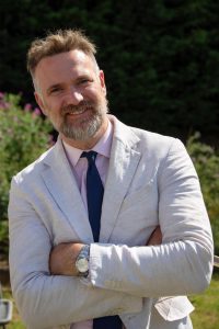 Charles Hanson, TV expert A man with grey beard and short hair, wearing a light jacket, pink shirt and blue tie. He's leaning a little to the left with his arms folded and smiling at the camera.