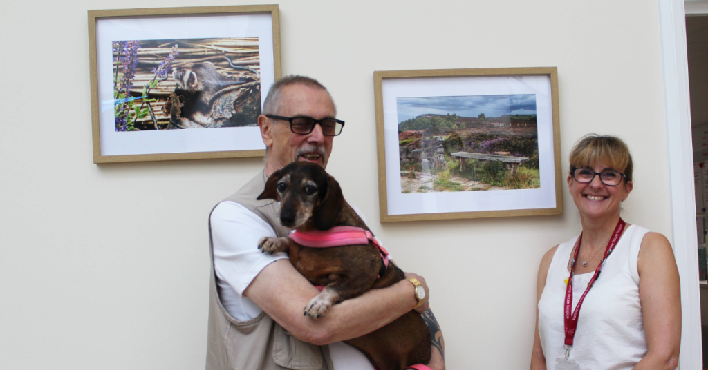 A man holding a small dog in front of two framed photos showing nature scenes that have been framed and mounted on a pale wall. Also standing with him is a lady smiling.