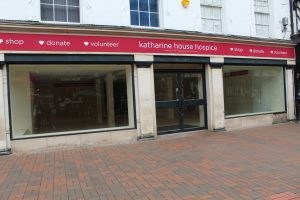 IMG_1556 A large building with two big windows that are empty and glass doors in the centre. A red sign above the windows and door states Katharine house hospice - shop - donate - volunteer