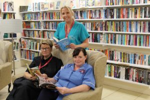 Staff enjoying some quiet time in the reading area before the opening Three ladies, two in nursing uniform. Two are sitting down on a sofa holding books and one is stood behind. Book shelves in the background full of books