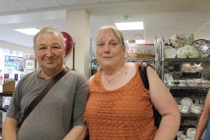 Coral and Paul An older man in a grey t-shirt smiling next to his wife in an orange standing in front of shelves stocked with crockery