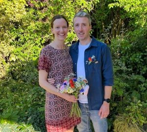 A couple standing together at their wedding blessing surrounded by greenery on a sunny day.