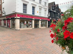 The new town centre location Stafford town centre showing some red flowers in the foreground and the new shop front. Above the door a red sign says 'Shop, volunteer, donate, Katharine House Hospice.