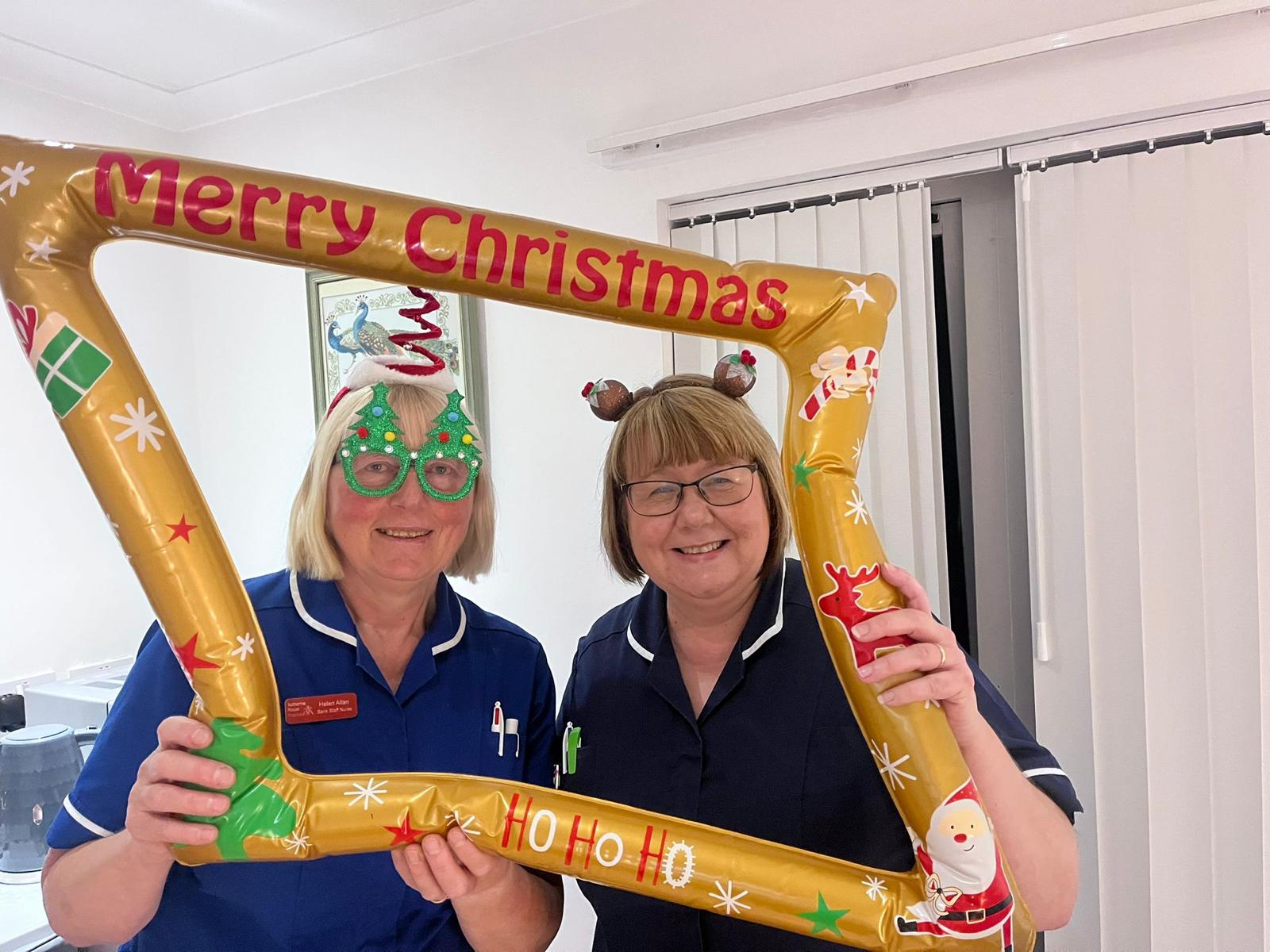 Two ladies in blue nurse uniforms with Christmas headbands and glasses holding an inflatable photo frame that says 'Merry Christmas'.
