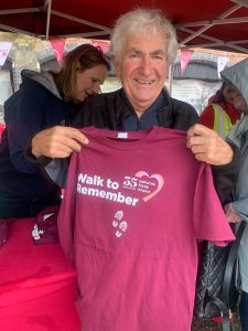 A man with grey hair holding up a maroon t-shirt with the Walk to Remember and Katharine House Hospice logos on it.
