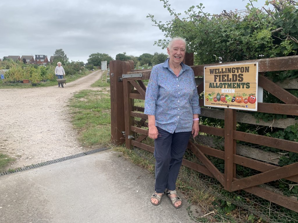 Women stands smiling by the entrance to allotments in country setting.