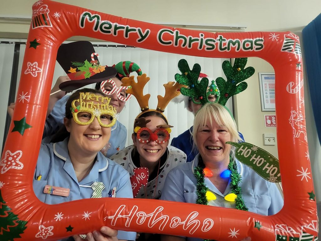 Five hospice nurses wearing Christmas novelty headbands with antlers and glasses while posing with an orange inflatable frame that reads Merry Christmas