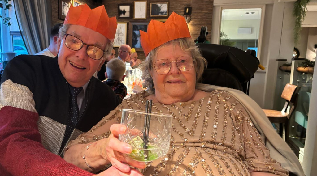 Carol with her husband sitting in a restaurant. Both wearing orange paper Christmas hats and Carol is holding a festive drink.