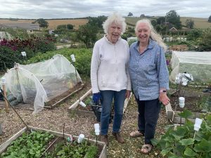 Two women standing side by side, smiling happily at their allotment garden in a rural countryside setting, surrounded by greenery and cultivated plots.