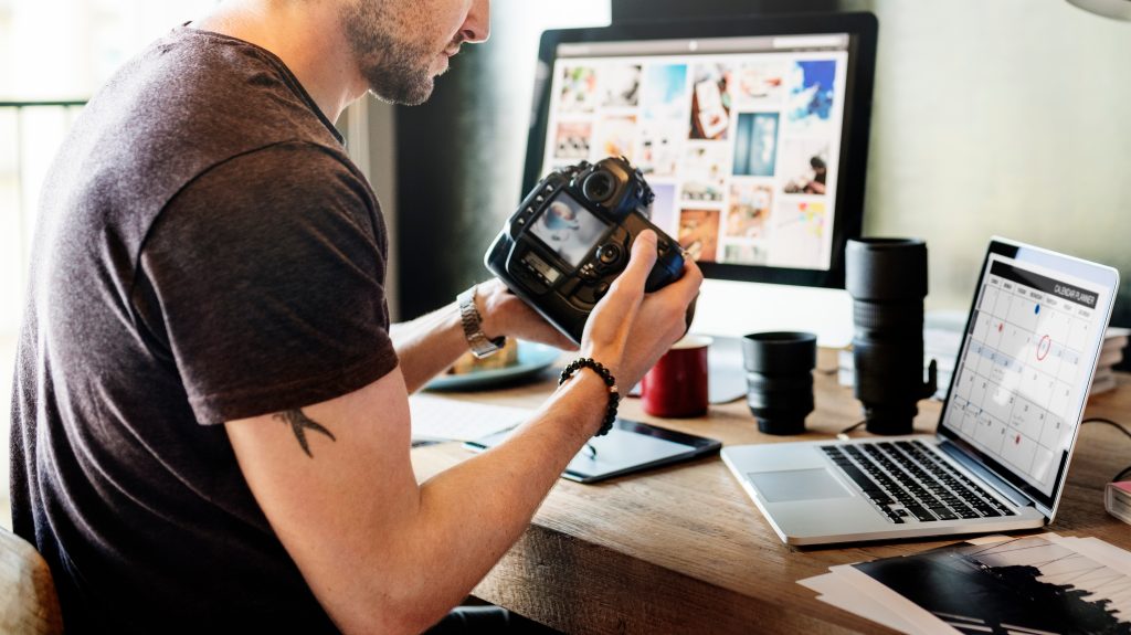 A man holding a camera. He is sitting at a desk in front of a laptop and has a partial tattoo showing on his arm