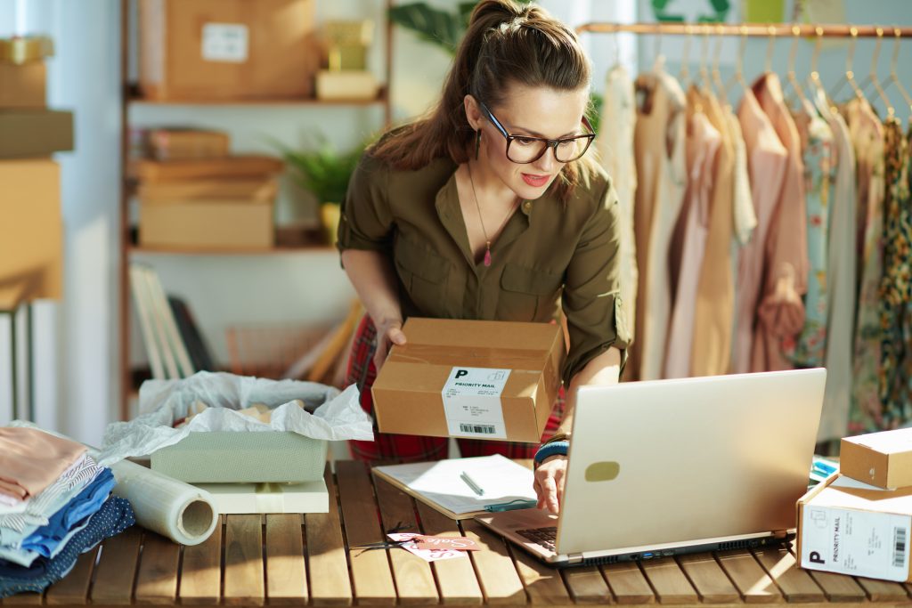 A young woman in a greenshirt holding a box and pressing keys on a laptop