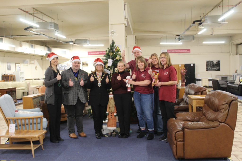 8 people wearing Santa hats and standing in front of a Christmas tree. There is a brown arm chair on the right and a wooden chair on the left.