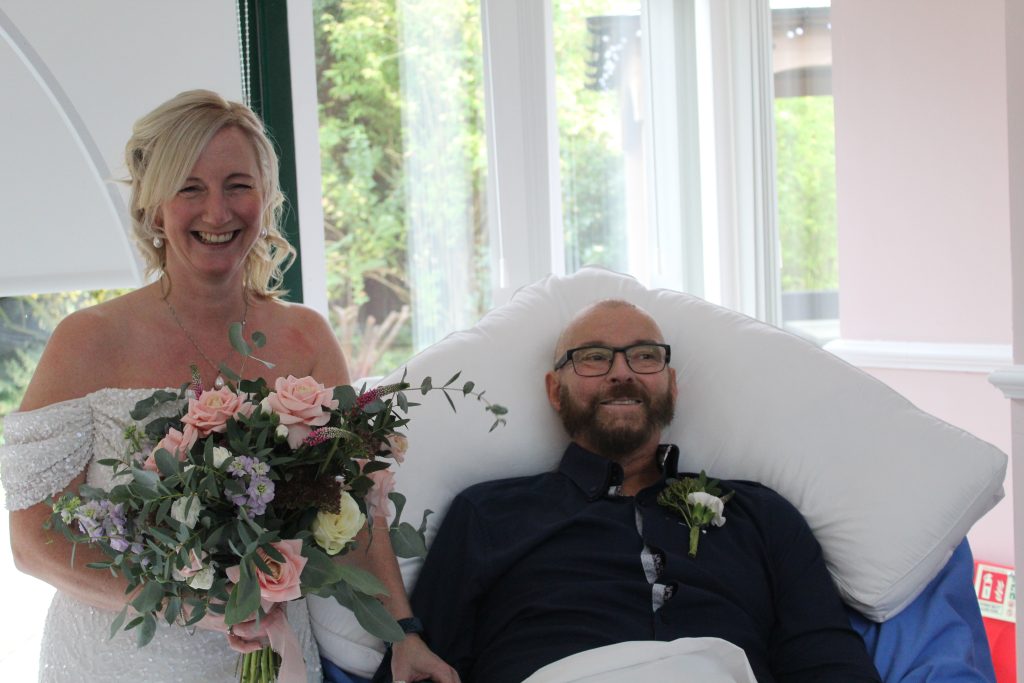 A bride wearing white holding a bouquet of white and pink flowers standing by the bedside of the goom who is sitting up wearing a dark shirt. Both are smiling.