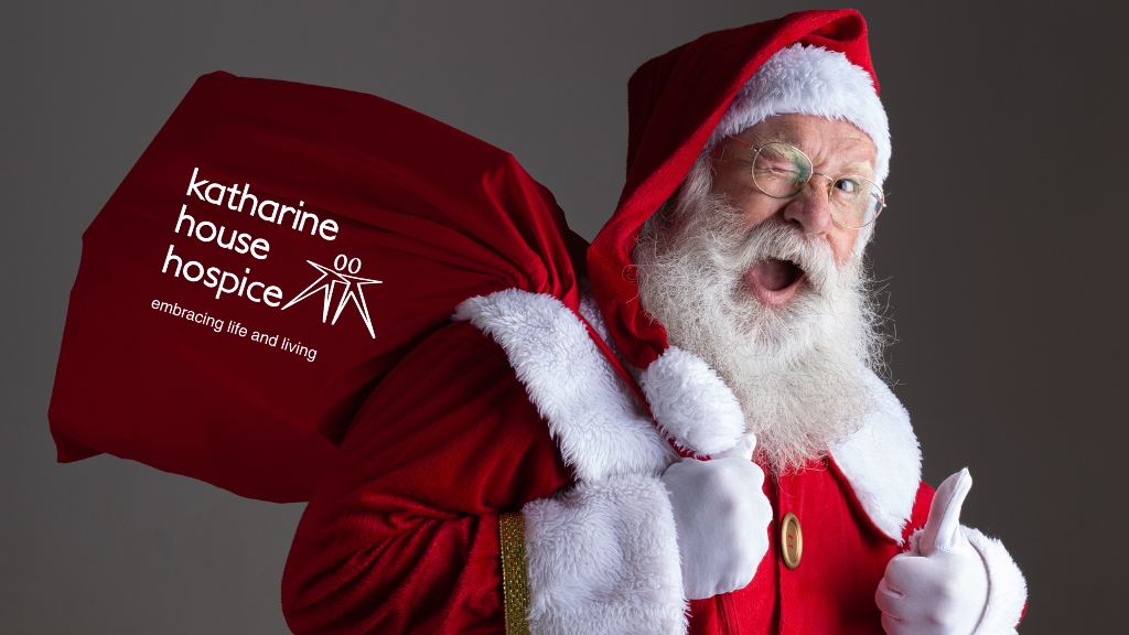 A white haired man with a white beard wearing a red santa suit and carrying a sack over his shoulder. He is winking and giving a thumbs up.