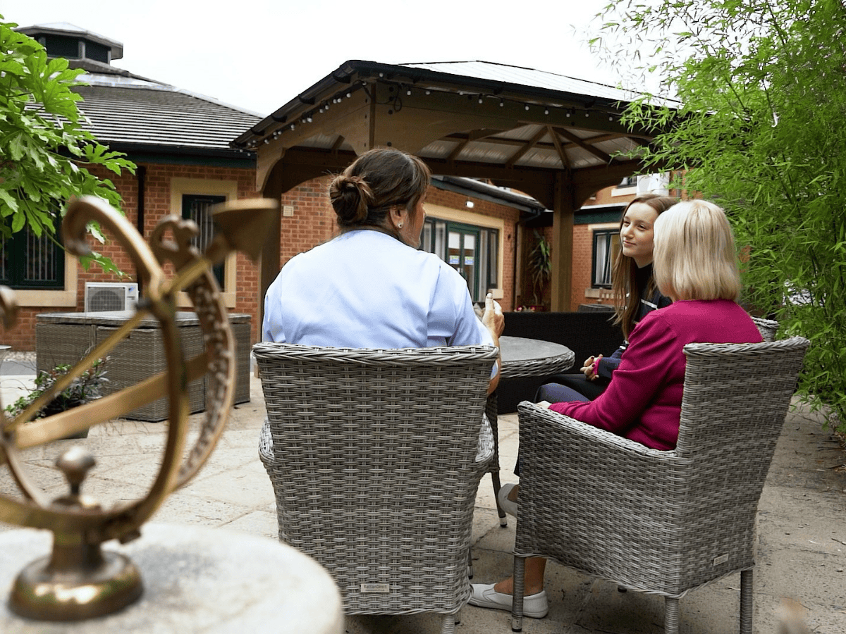 Chatting outside garden - 35 film (2) Three women sit in wicker chairs in a garden patio, engaged in conversation. A sundial and lush greenery create a peaceful scene.