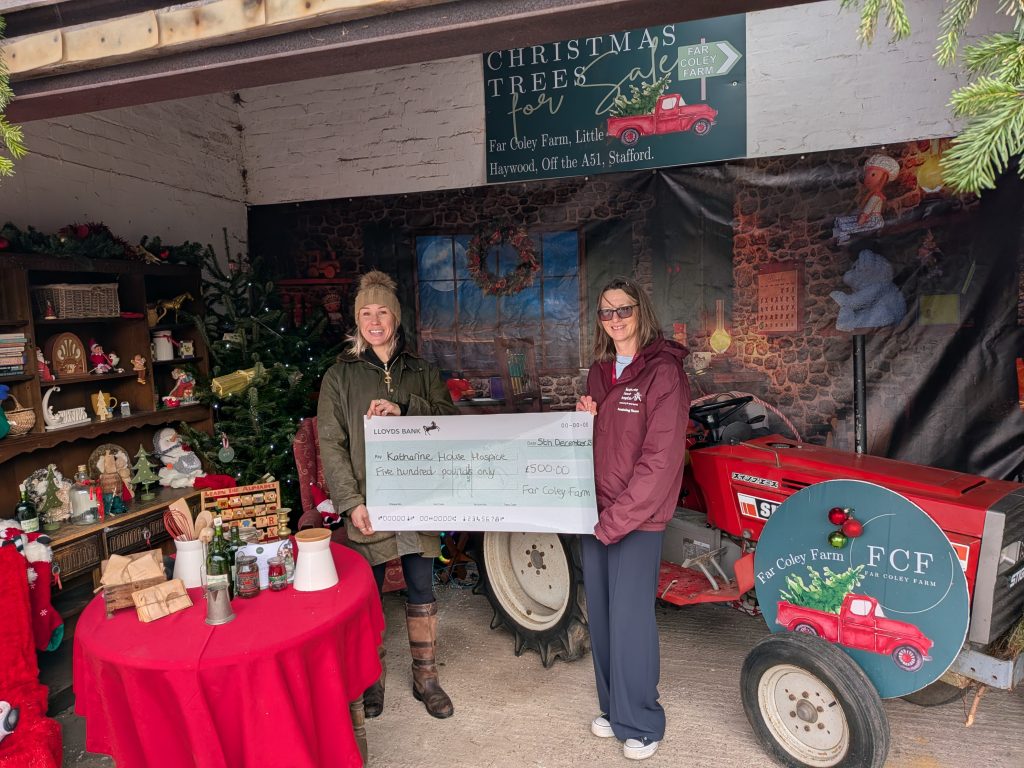 Tow ladies in a shed decorated with Christmas items. They are holding a giant cheque made out to Katharine House Hospice for £500