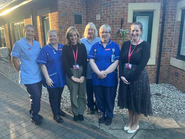 Three ladies in nurses uniforms and two other ladies in dark tops with lanyards standing outside a building
