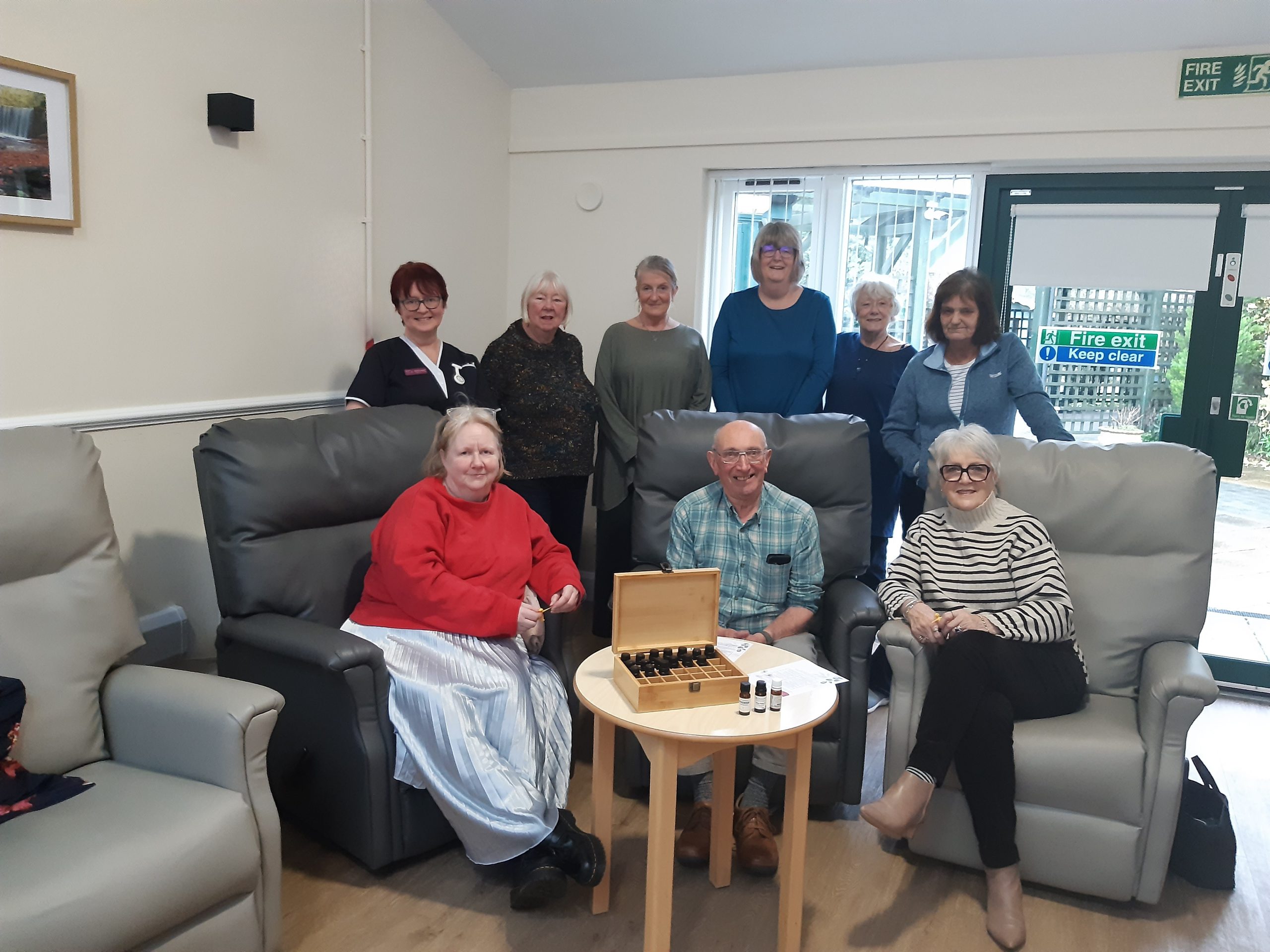 Carers and hospice staff gathered in a hospice lounge during a Wellbeing for Carers session. Some are seated in armchairs and others are standing behind them. A small table with relaxation oils is placed in the centre, reflecting the focus on wellbeing and support.