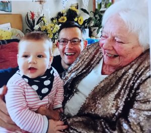 Older lady laughing with a smiling baby on her knee and a middle aged man steadying the baby and also laughing.