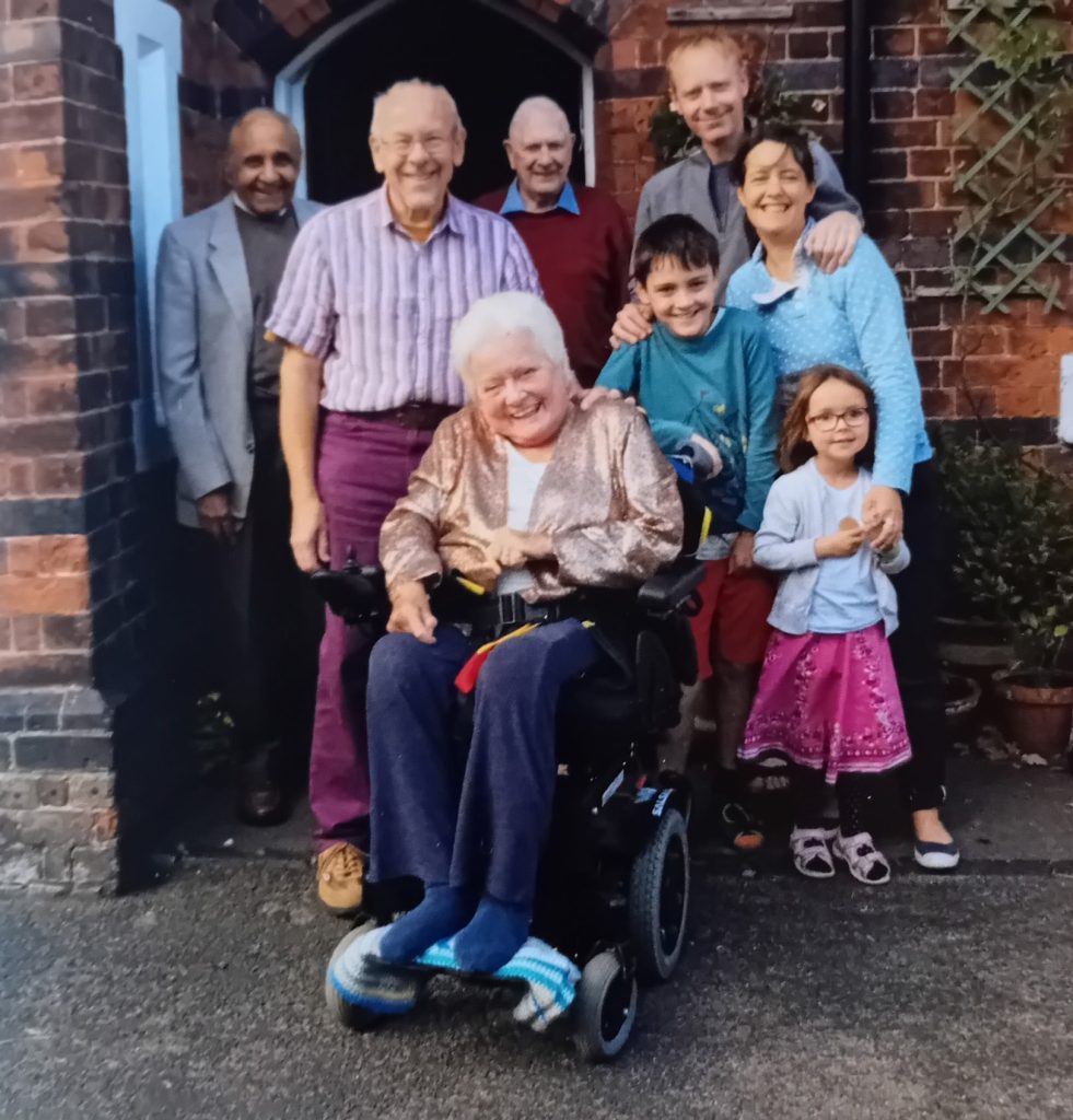 Val and family 2 crop Family group outside a front door including an older lady in a wheel chair, a couple with two young children and older men