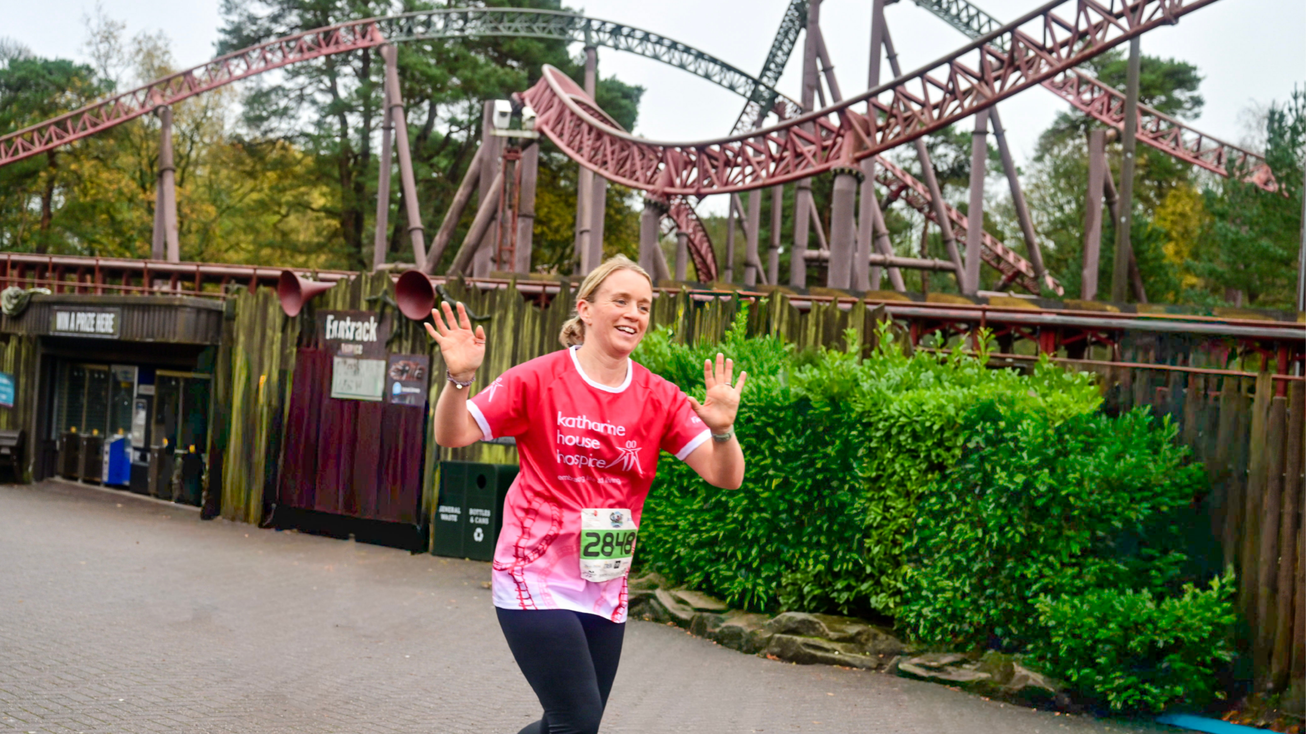 A woman wearing a Katharine House Hospice x Alton Towers top runs through the theme park, smiling and waving at the camera.