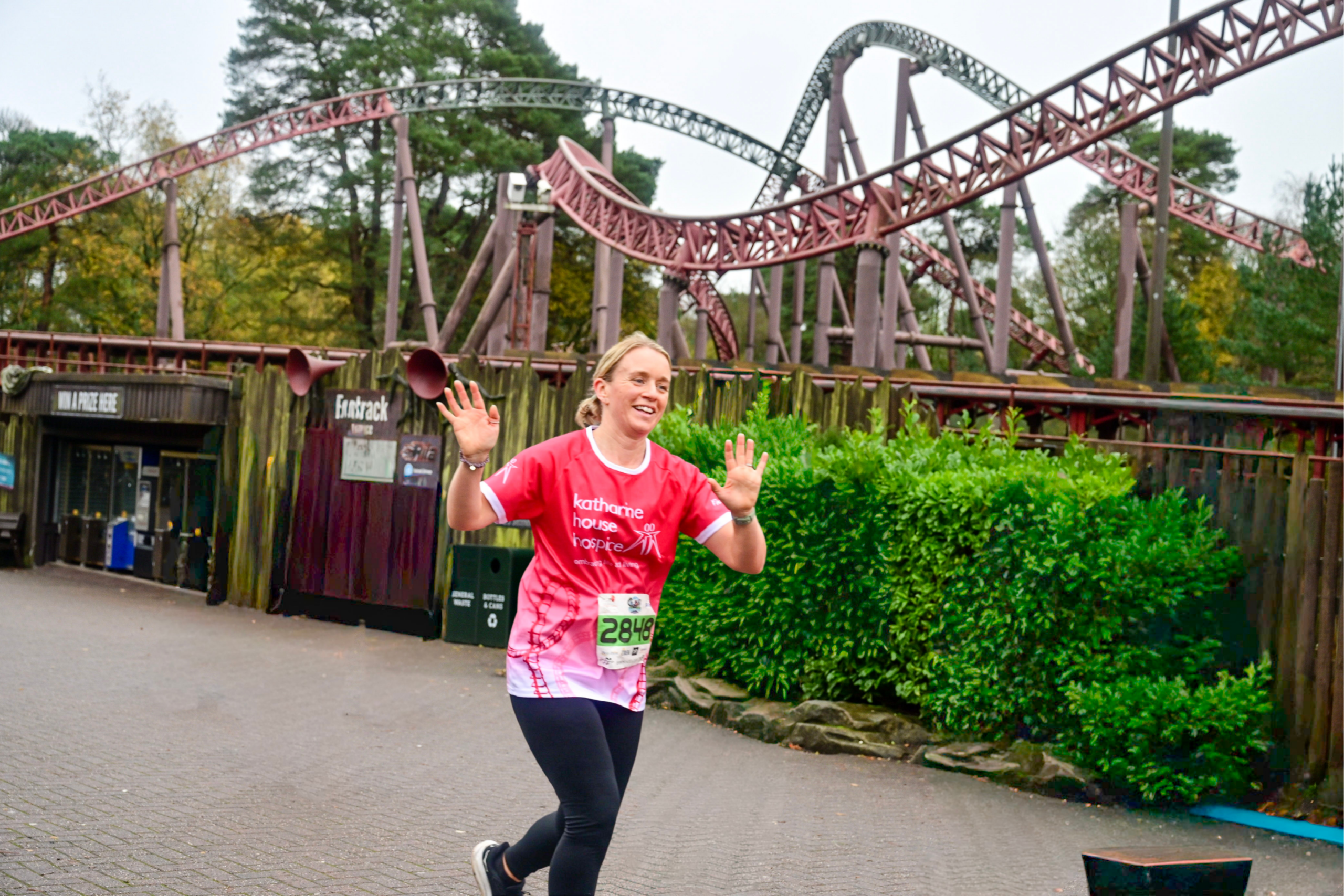 A woman wearing a Katharine House Hospice x Alton Towers top runs through the theme park, smiling and waving at the camera.