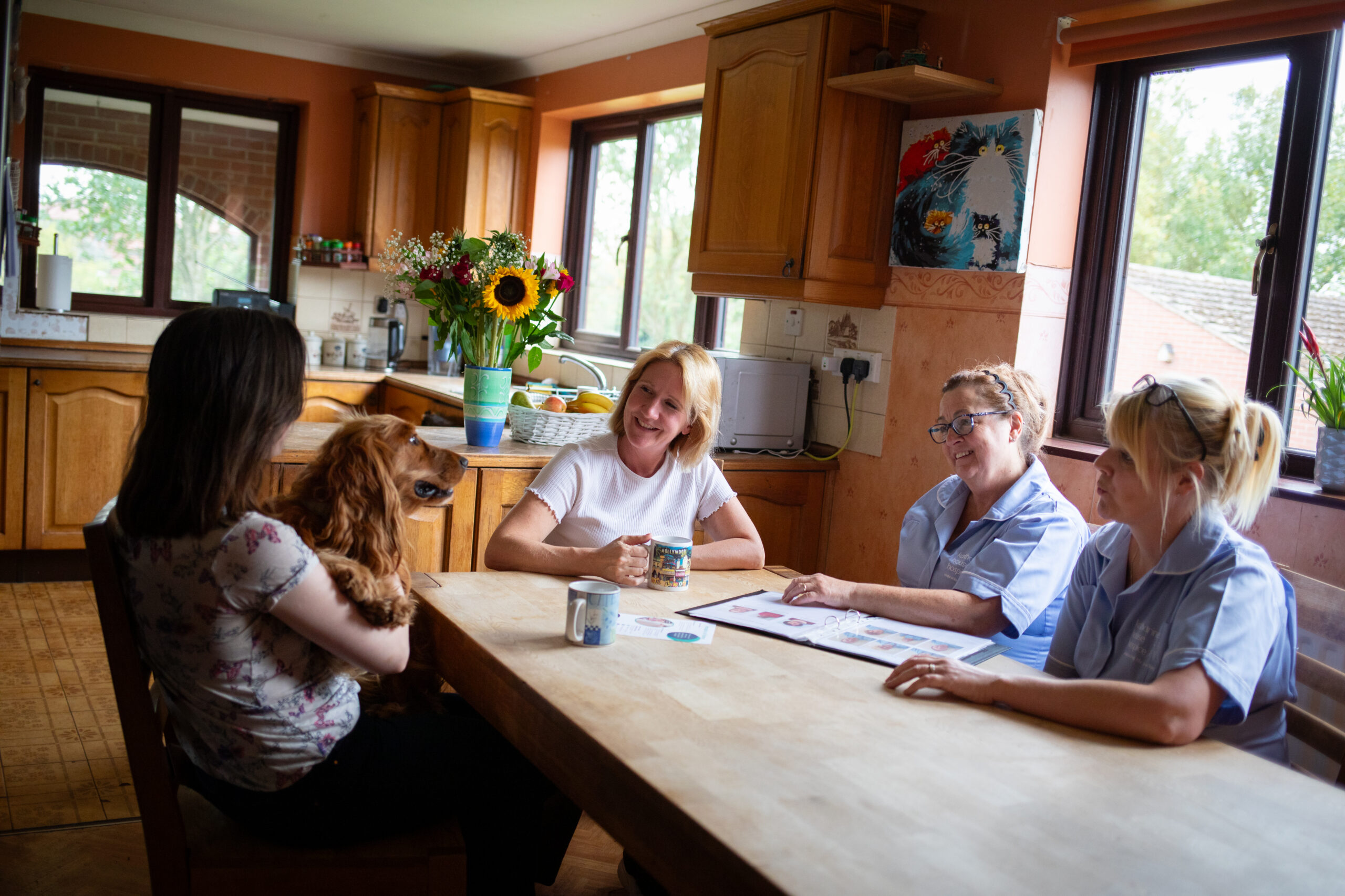 A woman, her daughter and two nurses sit at a kitchen table. A dog sits in the daughters lap, everyone smiling at the dog.