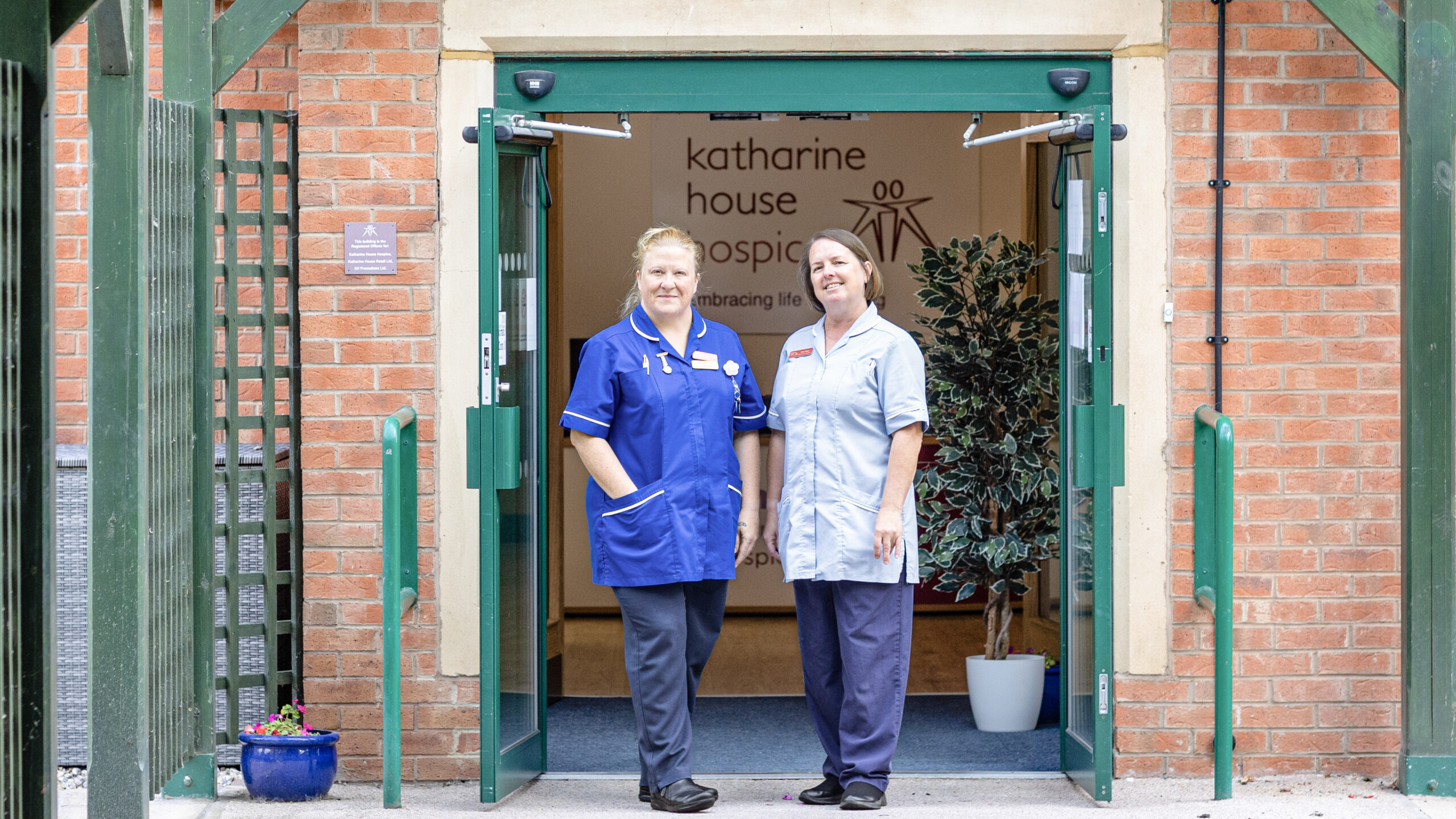 Two nurses stand at the open entrance of Katharine House Hospice, smiling.