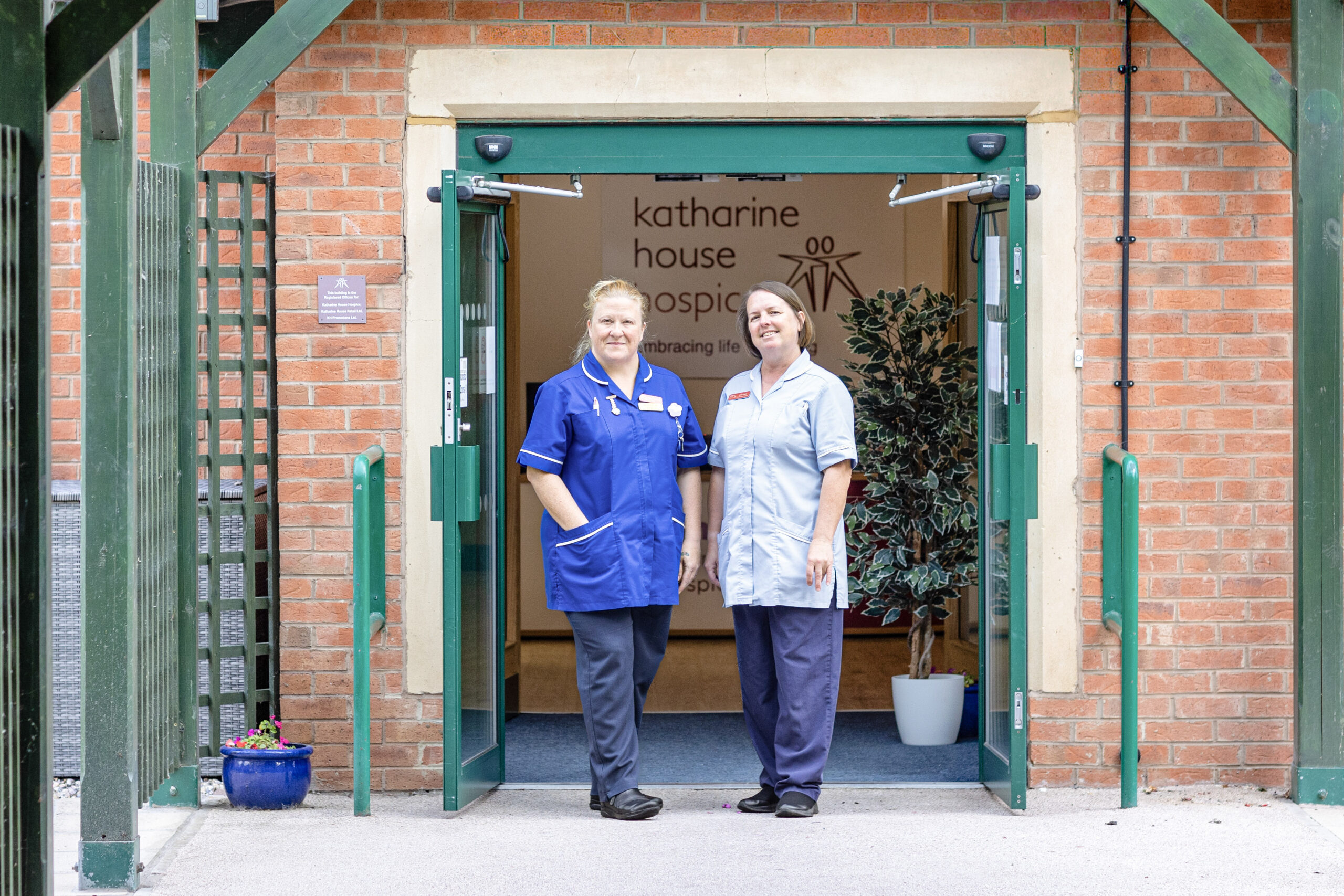 Two nurses stand at the open entrance of Katharine House Hospice, smiling.