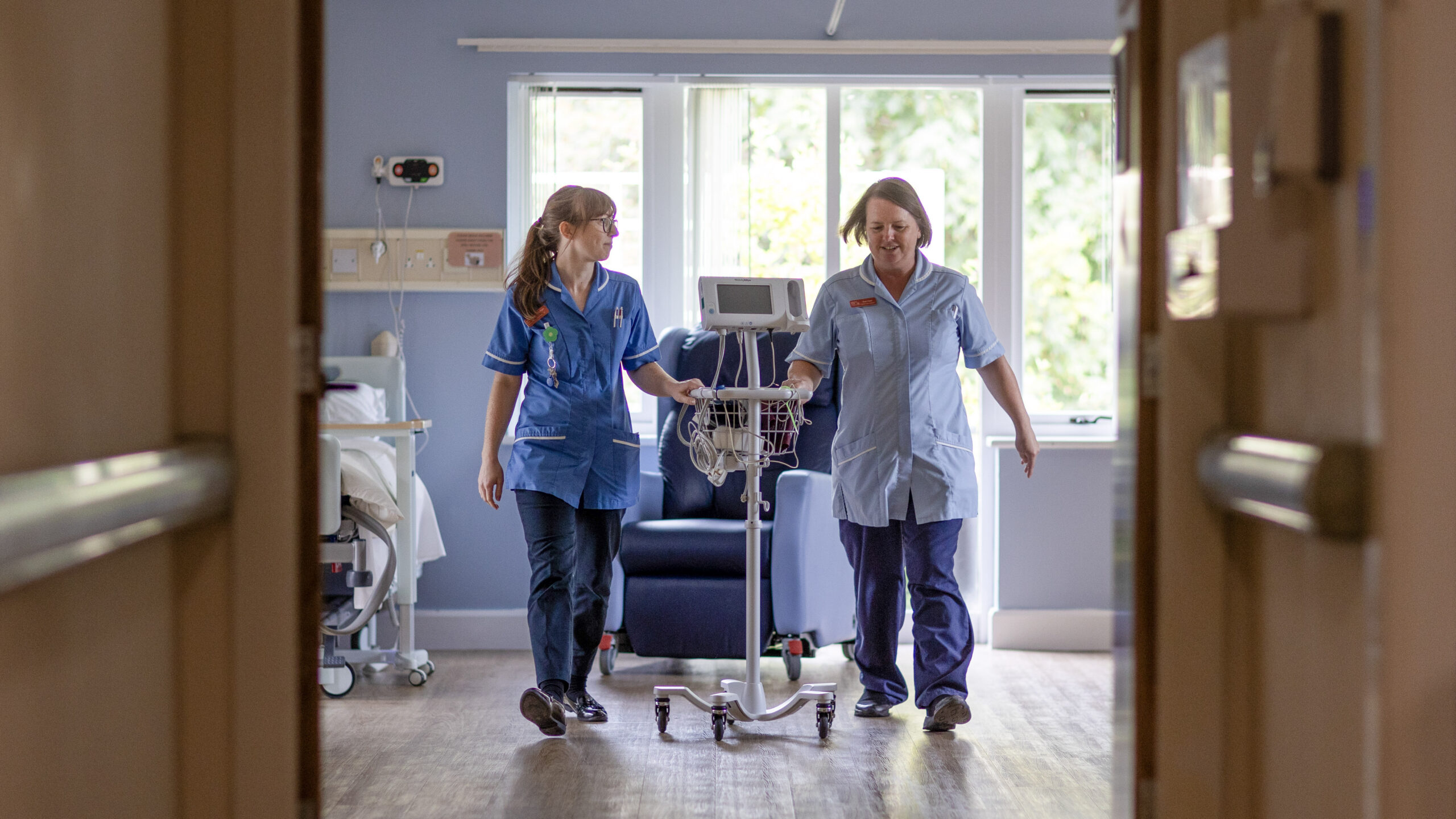 Two nurses wheeling a piece of equipment through a room.