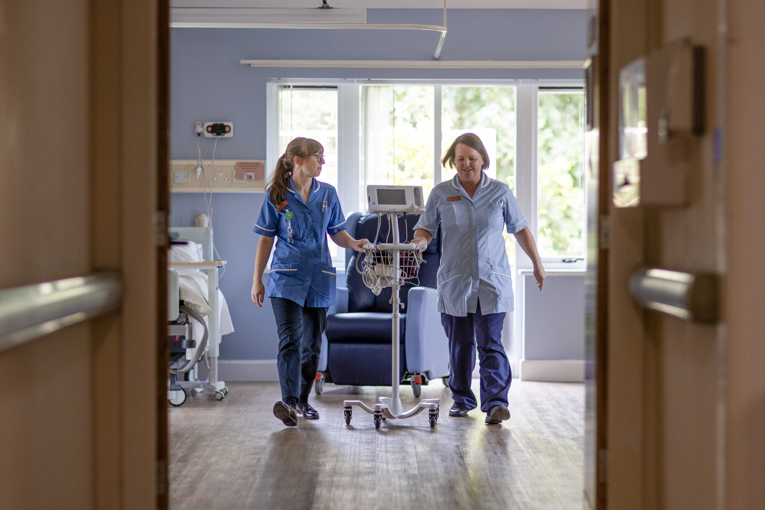 Two nurses wheeling a piece of equipment through a room.