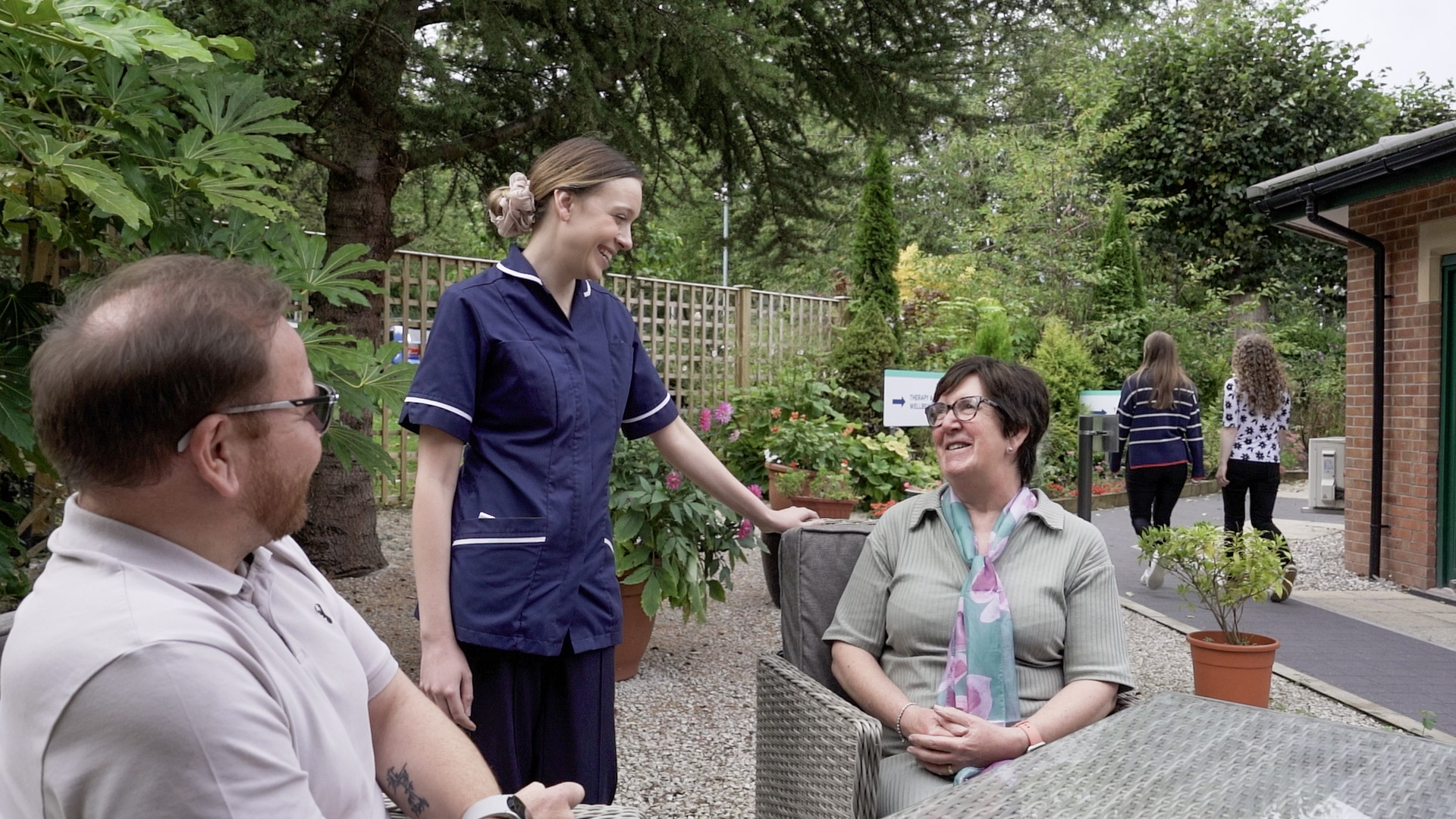 A nurse interacting with two people outside, surrounded by greenery.