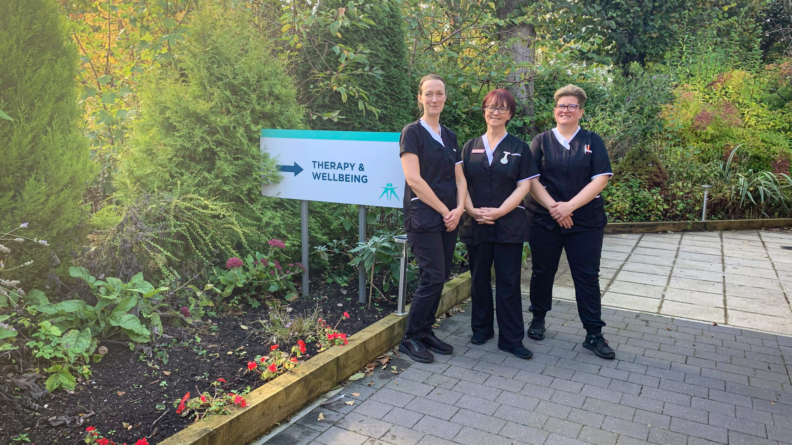 Three complementary therapists stand outside next to a 'therapy and wellbeing' sign and colourful plants.